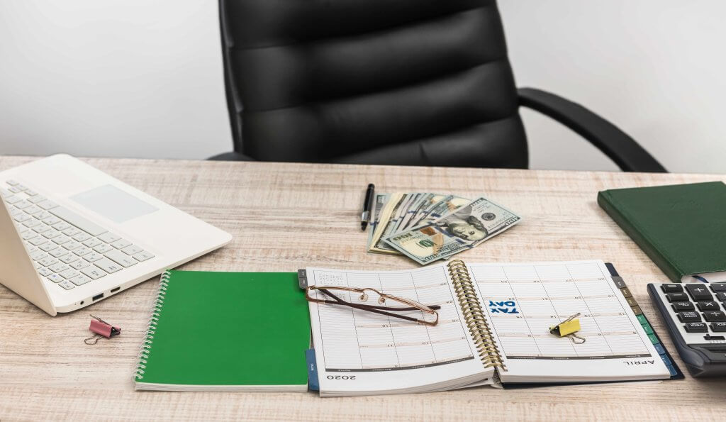 Desk with a monthly planner, calculator, cash, and laptop